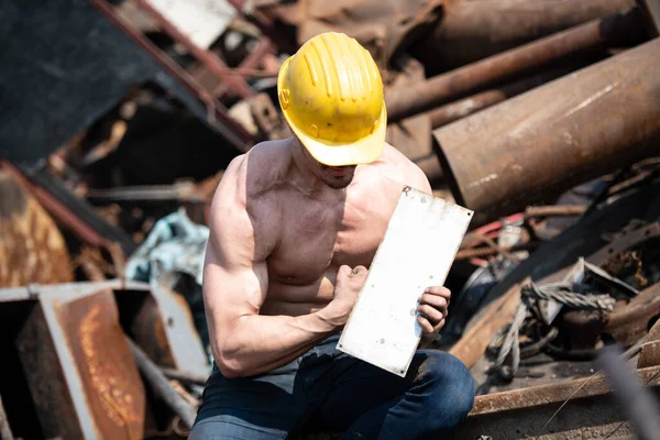 Portrait of a Young Physically Fit Man Sitting in Industrial Junk Yard Showing His Well Trained Body With Yellow Helmet