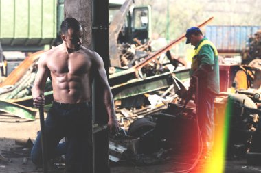 Endüstriyel hurdalıkta elinde kürekle duran ve iyi eğitilmiş vücudunu gösteren genç bir adamın portresi.
