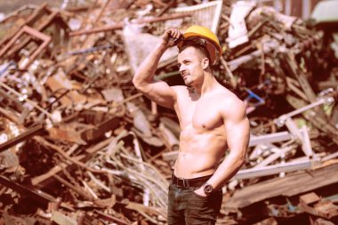 Endüstriyel hurdalıkta dikilen ve iyi eğitilmiş vücudunu sarı kaskla gösteren genç bir adamın portresi.