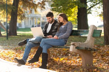 Young Couple Sitting on Bench in the Beautiful Autumn Day and Working On Laptop In Park