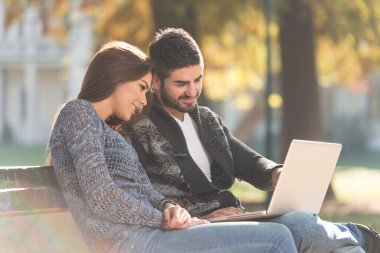 Young Couple Sitting on Bench in the Beautiful Autumn Day and Working On Laptop In Park
