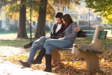 Young Couple Sitting on Bench in the Beautiful Autumn Day and Typing on the Phone In Park