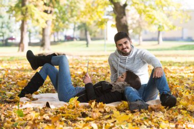 Güzel bir sonbahar gününde battaniyede oturan ve dijital tablet kullanan genç çift Park 'taki kulaklıkları dinliyor.