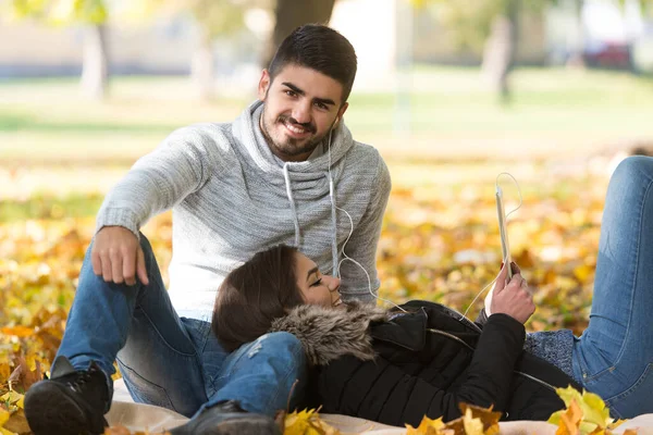 美丽的秋日里 一对年轻夫妇坐在毛毯上 在公园里用数码计时器聆听音乐 — 图库照片