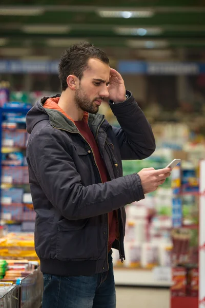 Hombre buscando confundido en el teléfono móvil en el supermercado —  Fotos de Stock