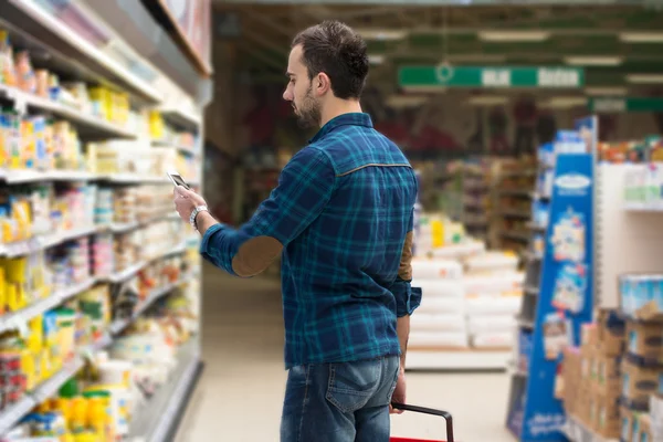 Młody człowiek wpisując na telefon komórkowy w supermarkecie — Zdjęcie stockowe