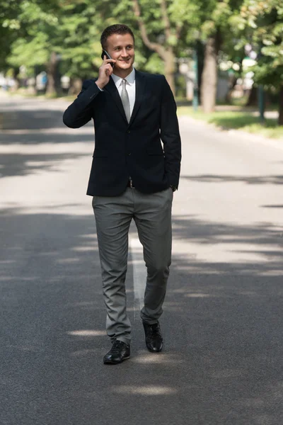 Empresario caminando al aire libre en el parque mientras usa el teléfono móvil —  Fotos de Stock