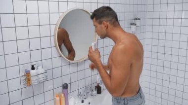 Tişörtsüz bir adam deodorant sıkarken banyoda sopa tutuyor.