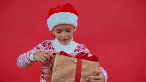 Mädchen Mit Weihnachtsmütze Und Weihnachtspullover Öffnen Geschenkbox Isoliert Auf Rot — Stockvideo