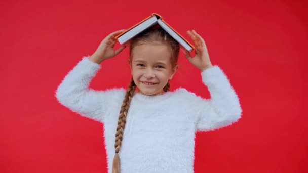 穿着蓬松毛衣的快乐女孩 把书放在头上 与红色隔离 — 图库视频影像