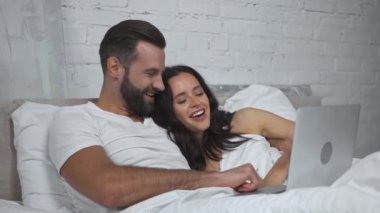 Mutlu çift yatakta dizüstü bilgisayarda film izlerken gülüyor.