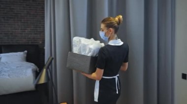 Tıbbi maskeli genç oda hizmetçisi temiz havlularla dolu bir kutu taşıyor. 