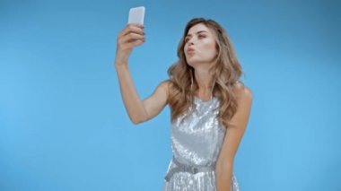 Elbiseli kadın akıllı telefondan selfie çekiyor, mavi renkte izole edilmiş. 