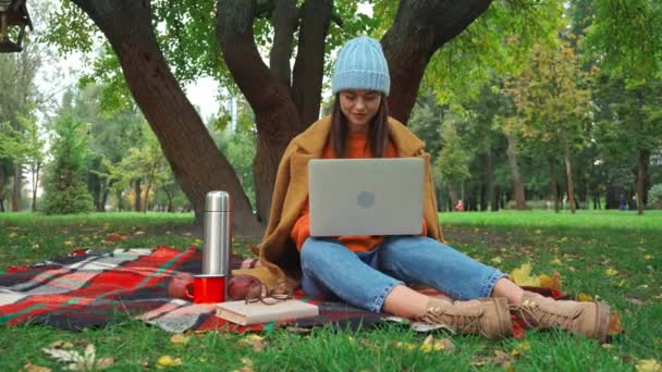 穿着秋季服装的自由职业者在公园的格子毛毯上在笔记本电脑上打字 — 图库视频影像