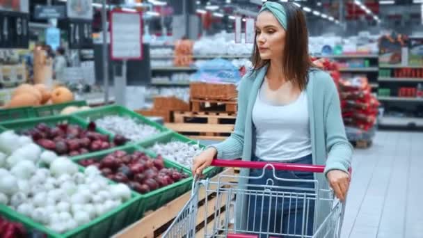 在超级市场 一个年轻女子提着购物车走在模糊的蔬菜旁 — 图库视频影像
