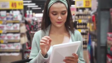 Genç kadın süpermarkette yürürken dijital tablette listeyi kontrol ediyor