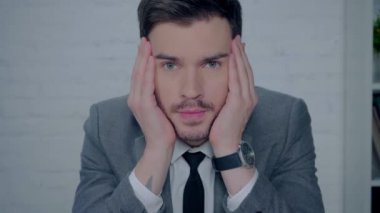 Sıkılmış iş adamı yanaklarına dokunuyor ve kameraya bakıyor.