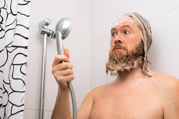 满脸胡须 头晕的男人站在浴室里 看着淋浴 而供水却停止了 水龙头没水喝 — 图库照片