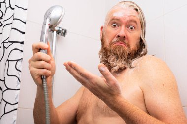 Bilmece gibi ıslak sakallı adam elinde duşla küvette duruyor ve şaşkınlıkla kameraya bakıyor. Su kesme konsepti