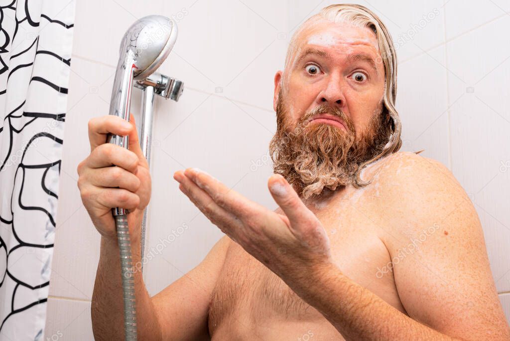 Puzzled wet soapy bearded man standing in bathtub holding shower in his hand and looking at the camera in bewilderment. Water cut-off concept