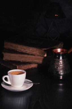 White cup of fresh hot coffee, copper cezve and stack of old books on black wooden table in the dark. Low key. Copy space