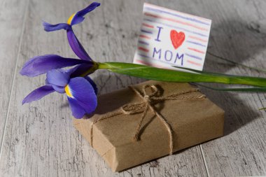 Kraft kağıdıyla sarılı bir hediye kutusu, üzerinde anne ve Iris 'e sabah güneşinde gri ahşap masada mesaj olan bir çocuk kartı. Anneler Günü tatili konsepti