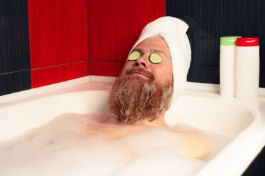 Havluya sarılmış kafa ve gözlerinde salatalık dilimleriyle banyo yapan sevimli sakallı adam. Komik hipster köpüklü küvette rahatlıyor ve hayattan zevk alıyor. SPA evde.