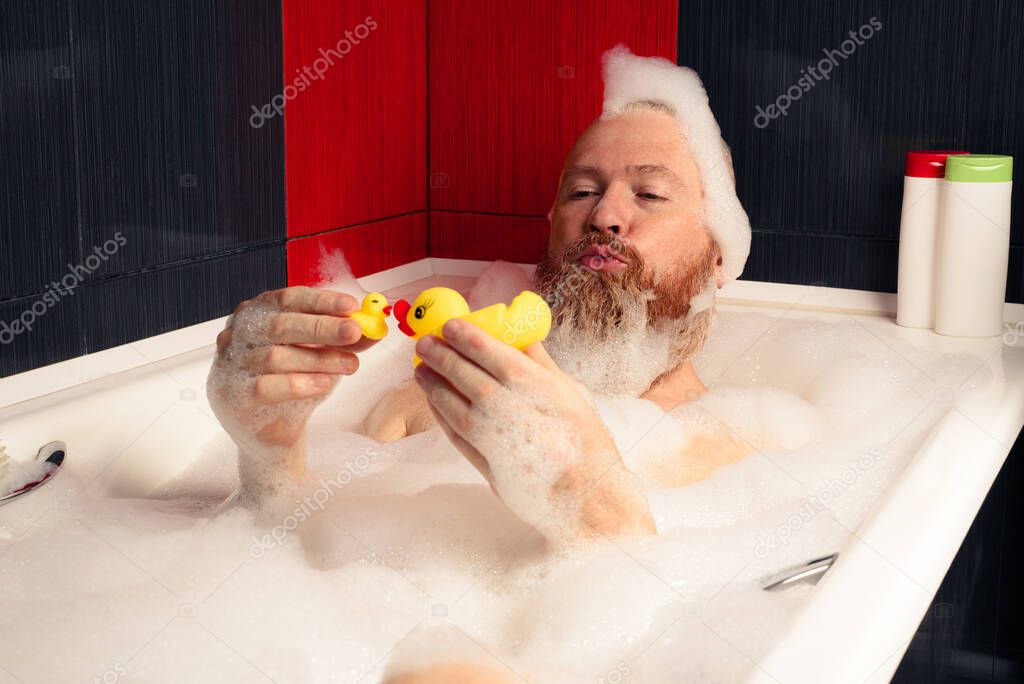 Cute funny bearded man playing with rubber ducks in bubble bath relaxing at home.