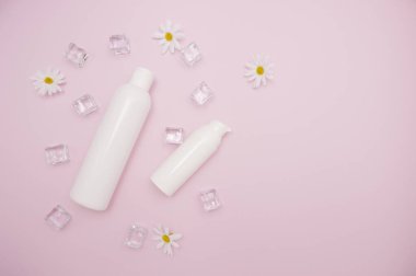 Body and skin care, cosmetology. White plastic bottle for shampoo and other cosmetics. Ice cubes around, daisies, pink background, free space for text, top view.