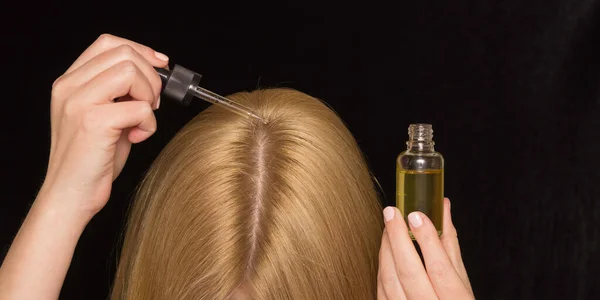 Women's hair and hands with a cosmetic oil and a pipette. Care for the scalp, healthy hair. Black background.