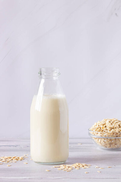 Овсяное молоко в стеклянной бутылке и овсяные хлопья на светлом фоне. Здоровые напитки, вегетарианство. Вид сбоку. Вертикальное фото