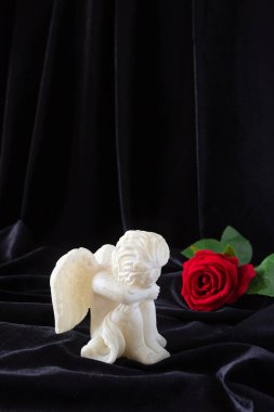 A small sculpture of a baby angel with wings and a red rose on a black background. The concept of funerals, condolences, and mourning. clipart