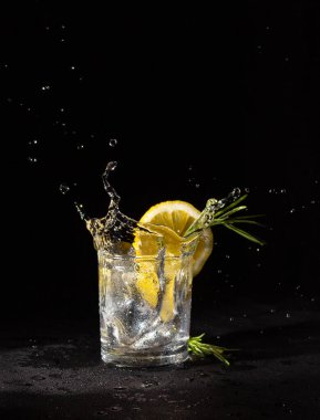 Colorless alcoholic drink in a glass with a slice of lemon and rosemary, splash. Vodka on a black background. clipart