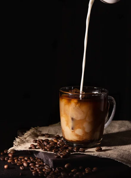 Glass cup with cold coffee and ice, coffee beans on a dark background. A hand pours cream.