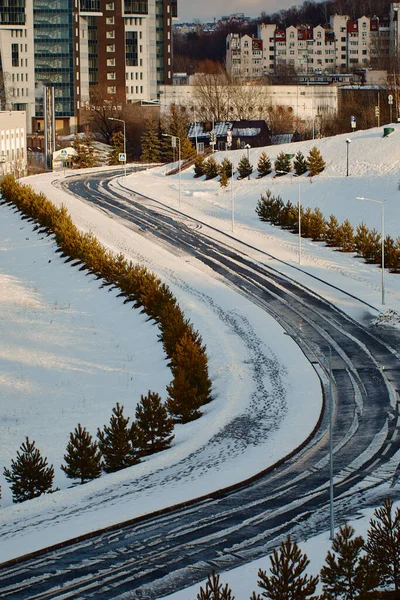 Curved road with tire tracks in the snow leading to a group of houses. Trees grow on the sides of the road. Kazan, Russia