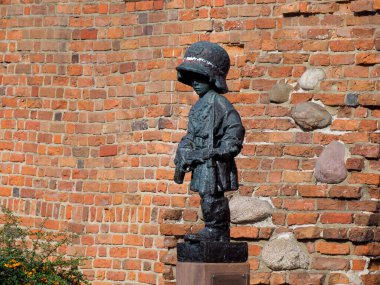 Küçük, koyu bronz bir kasklı bir çocuk heykeli eski bir kırmızı tuğla duvarın dokulu arka planına karşı.