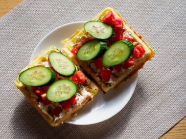 Krem peynirli iki iştah açıcı waffle, dilimlenmiş domates, ve beyaz bir tabakta taze salatalık, dokuma hasırında servis edilir..