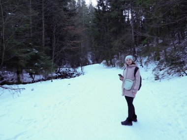 Kışın karlı bir ormanda yürürken elinde telefon tutan pembe paltolu genç bir kadın..
