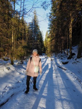 Kışın giyinmiş genç bir kadın güneşli bir çam ormanında karlı bir yolda yürüyor.