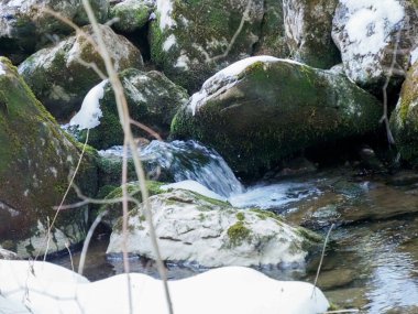 Kristal berrak su, kış mevsiminde büyük yosunlu kayaların üzerine düşüp karlı bir dere yatağına düşüyor.