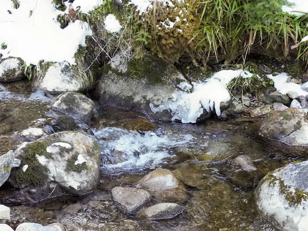 Dağdaki bir derede eriyen buzun ve akan suyun karanlık taşların üzerinden detaylı görüntüsü.