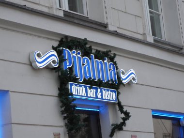 Beyaz duvarda yeşil çam çelenkleriyle süslenmiş Pijalnia barının neon mavisi tabelası.