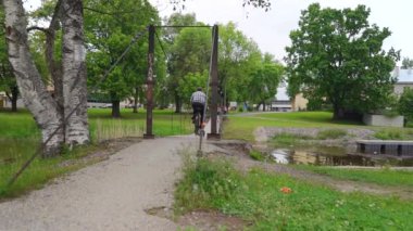 Yaşlı bir adam nehrin karşısındaki asma köprüde bisiklet sürüyor. Yavaş çekim görüntüleri.