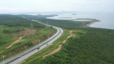 Ormanlar ve tarlalar arasında arabalar yol boyunca ilerler. Deniz kıyısı ufukta görünüyor..