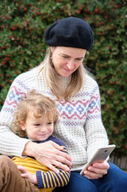 Küçük şirin bir çocuk annesiyle bankta oturuyor ve cep telefonuna bakıyor.