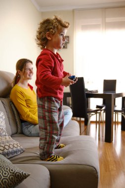 Çocuk video oyunu oynuyor, anne kanepeden izliyor.