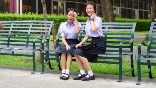 Söt asiatiska thailändska höga skolflickor student paret i skolan uniform sitter på en bänk som visar ett roligt leende medan hårda vinden blåser — Stockvideo