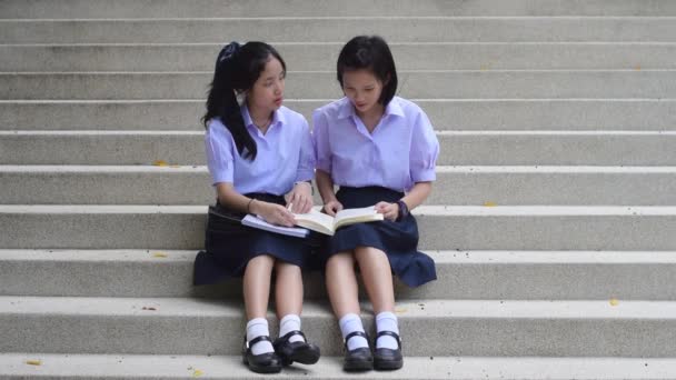 Cute Asian Thai high schoolgirls student couple in school uniform sit on the stairway discussing and reading homework or exam together. — Stock Video