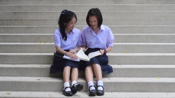 Lindo asiático tailandés colegialas secundaria estudiante pareja en uniforme escolar sentarse en la escalera discutir y leer la tarea o examen juntos . — Vídeo de stock