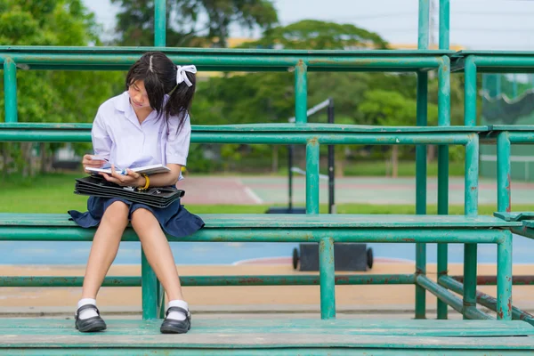 Lindo asiático tailandés colegiala estudiante en uniforme es sentado y leyendo en un metal stand lectura libros en escuela patio de recreo —  Fotos de Stock